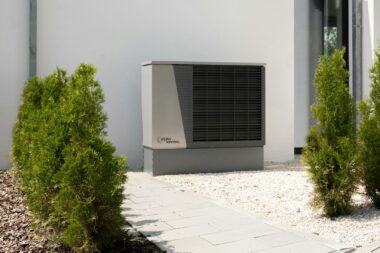 Outdoor air-source heat pump unit beside a modern white house with shrubs