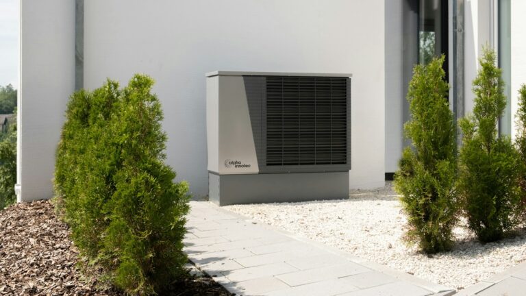 Outdoor air-source heat pump unit beside a modern white house with shrubs