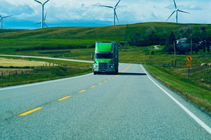 Green semi truck driving on a rural highway past wind turbines