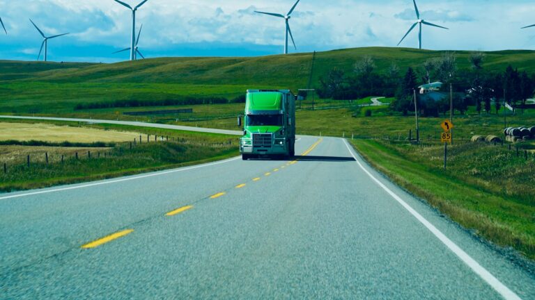 Green semi truck driving on a rural highway past wind turbines